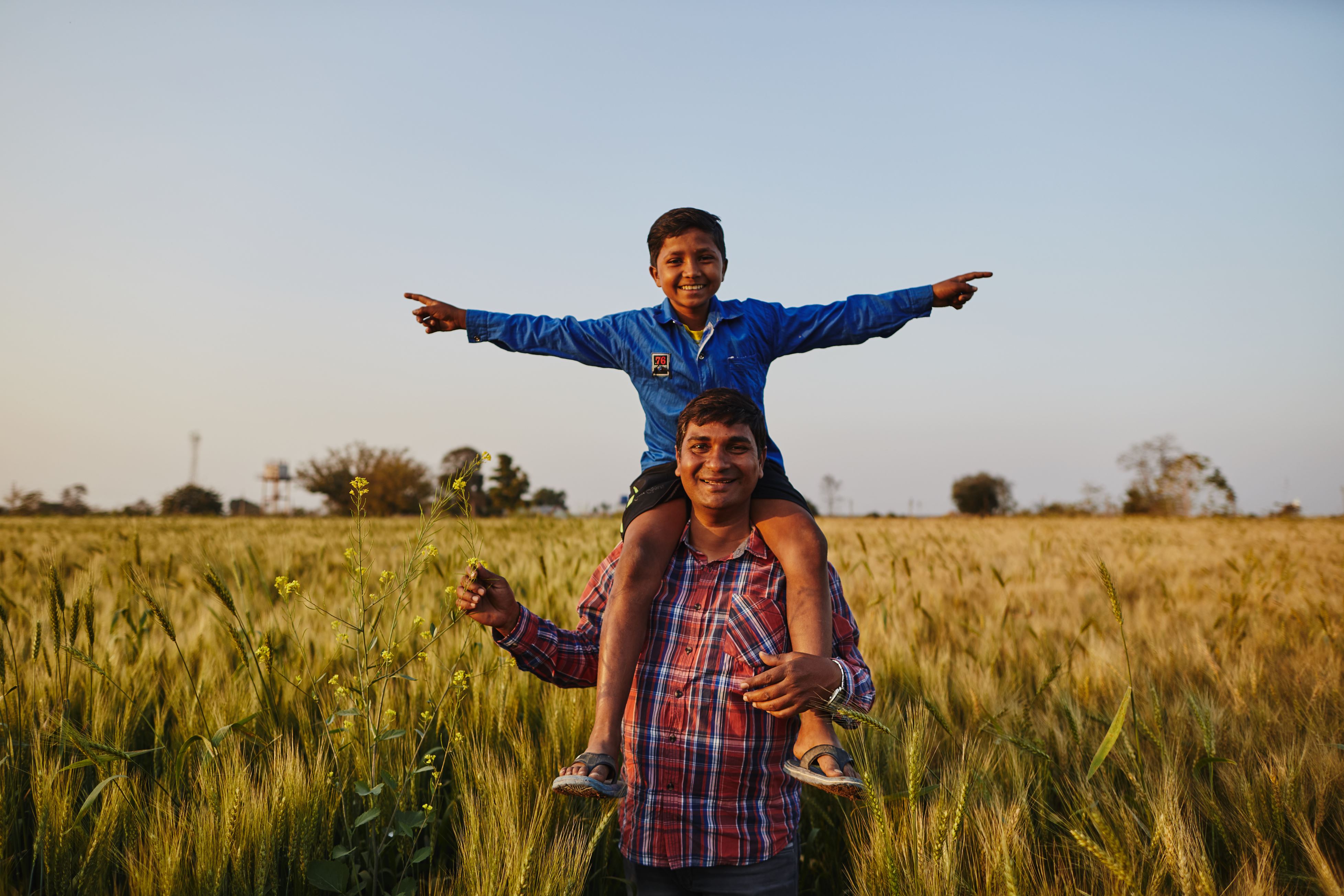 Bambino sostenuto a distanza sulle spalle del padre. Sorridono in un campo di grano. 