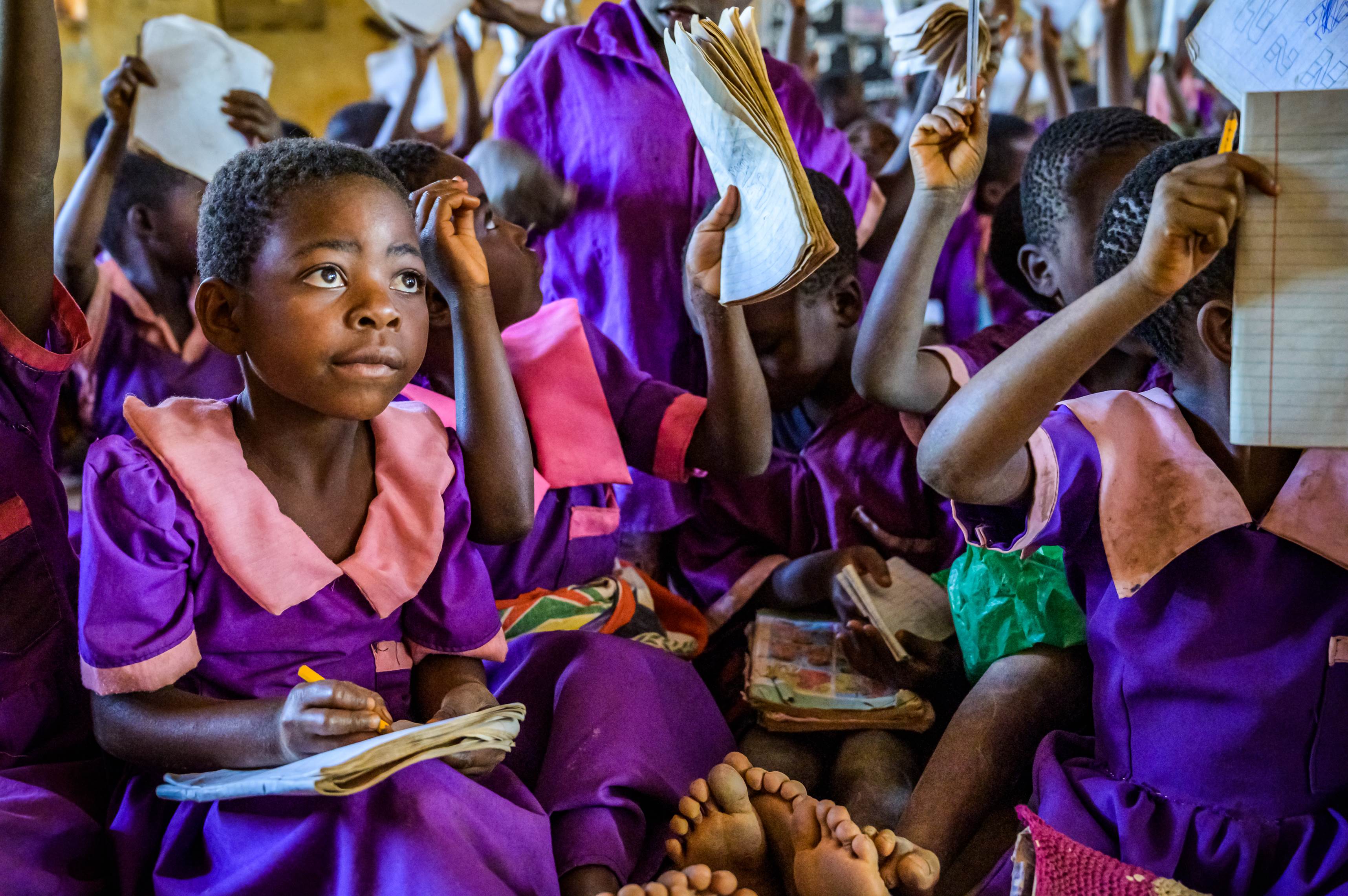 Una ragazza del Malawi in uniforme scolastica con un taccuino in grembo che ascolta la classe, con molti compagni di classe dietro di lei