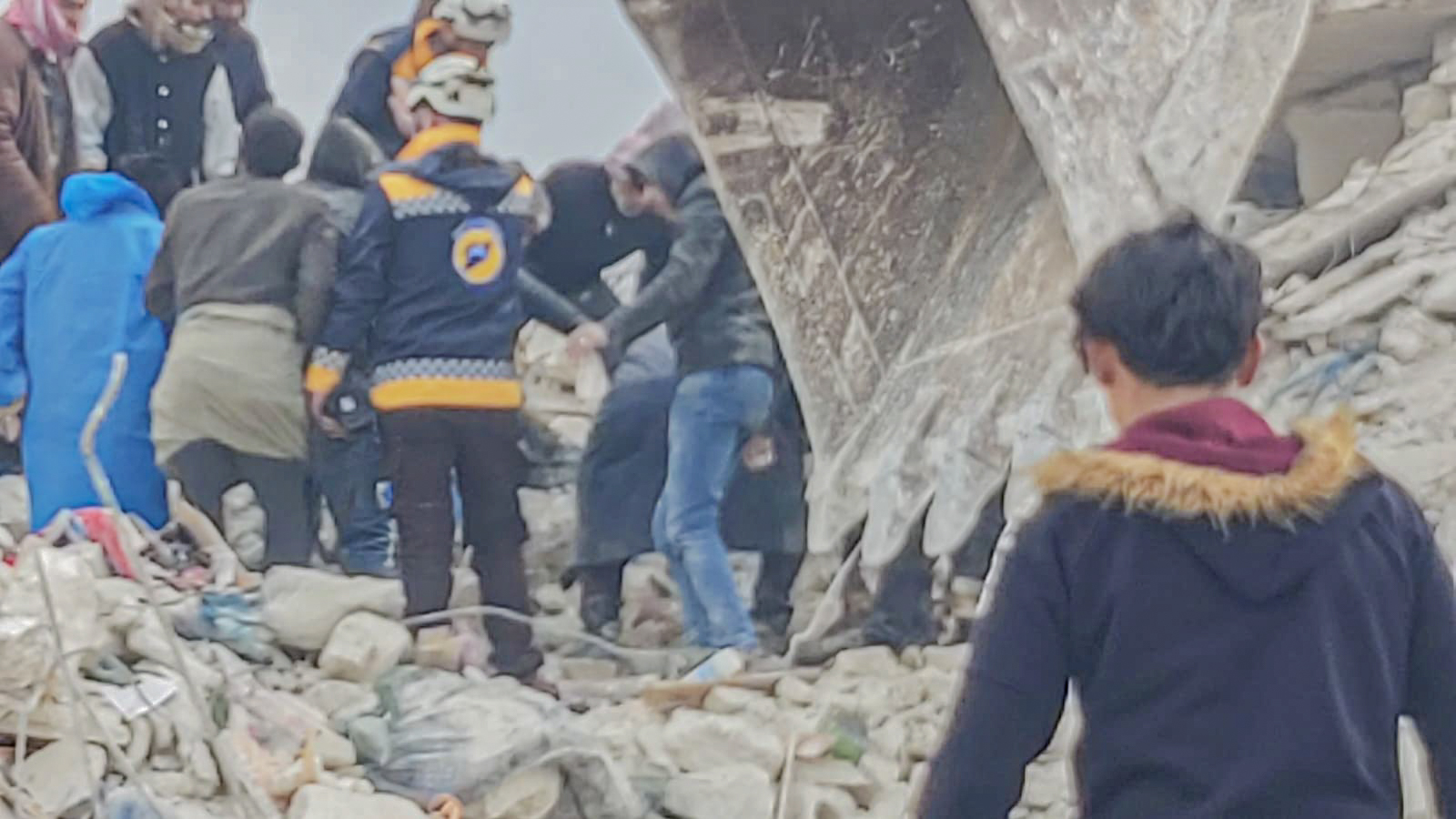 Un ragazzo con le spalle alla telecamera cammina verso un gruppo di persone che cercano di salvare altre persone da sotto le macerie dopo il terremoto in Siria