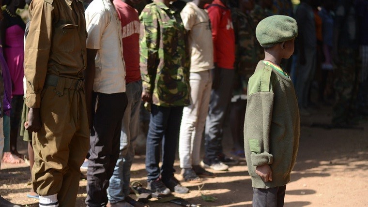 Persone in fila dietro un bambino che indossa una tuta mimetica militare e un berretto in Sud Sudan