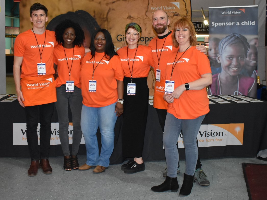 Sei dipendenti di World Vision in magliette arancioni davanti a uno stand che si offrono volontari a un evento World Vision