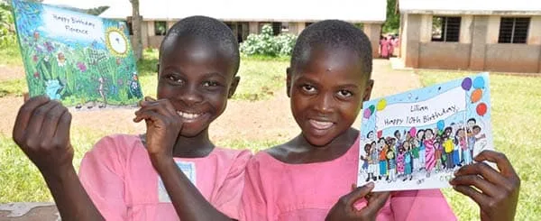 Due bambine africane sorridenti mostrano orgogliosamente dei biglietti di auguri colorati, all'aperto, indossando uniformi scolastiche rosa