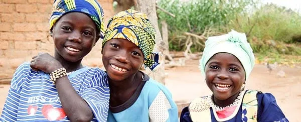 Tre bambine africane sorridenti, vestite con abiti colorati tradizionali e foulard, posano all'aperto davanti a un muro di mattoni e vegetazione