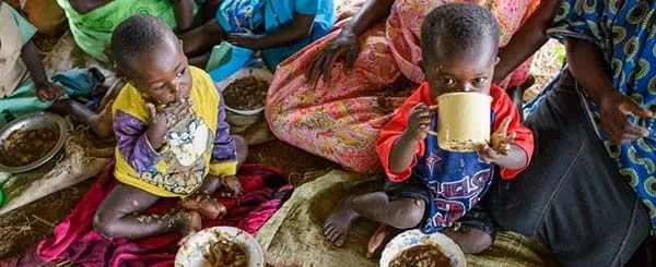 Due bambini piccoli seduti a terra mangiano da ciotole, uno beve da una tazza in un contesto familiare