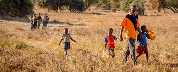 Adulto e bambini camminano in un campo erboso in un'area rurale, seguiti da altri membri della comunità